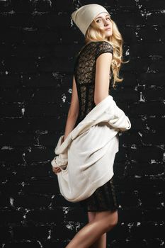 Fashion portrait of trendy girl with blond hair, wearing a black dress and jacket standing against the black urban wall. Street style for young lady. Youth look, hip hop and hipster trends
