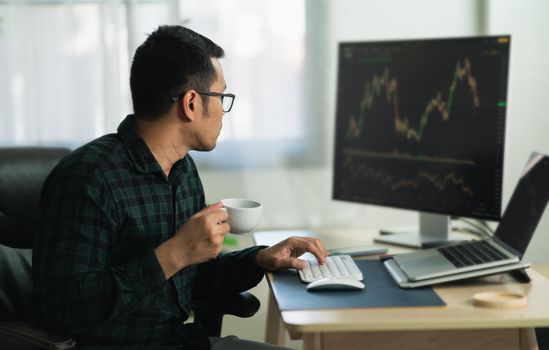 Asian man trader drinking coffee and sitting at home office in front of monitors with cryptocurrency graph holding smartphone monitoring cryptocurrency price