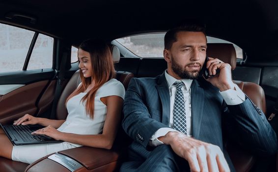 Young businesswoman and businessman sitting on the back seat of the car using mobile phones