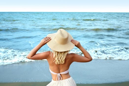 Blonde Woman in Fashion Summer Style Standing at Sea and Holding Hat. Luxury Lifestyle Rear View. Summer Vacation Concept with Ocean. Soul Relaxing, Spirit Calm