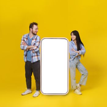Asian girl and caucasian guy standing leaned on huge smartphone with white blank screen, mobile app advertisement and smile looking at each other isolated on yellow background. Product placement.