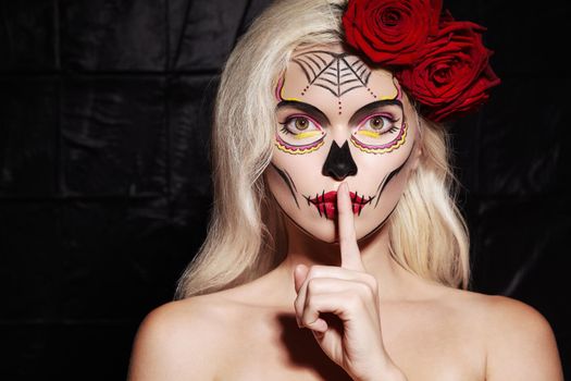Beautiful Halloween Make-Up Style. Blond Model Wear Sugar Skull Makeup with Red Roses, pale Skin Tones and Waves Hair. Santa Muerte concept. Silence Gesture