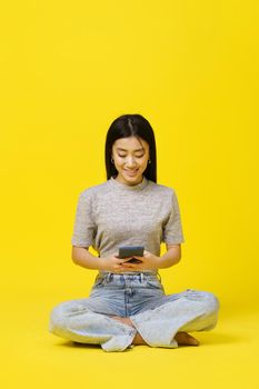 Asian young girl sitting on the floor holding smartphone texting or shopping online isolated on yellow background. Product placement for mobile app advertisement. Copy space.