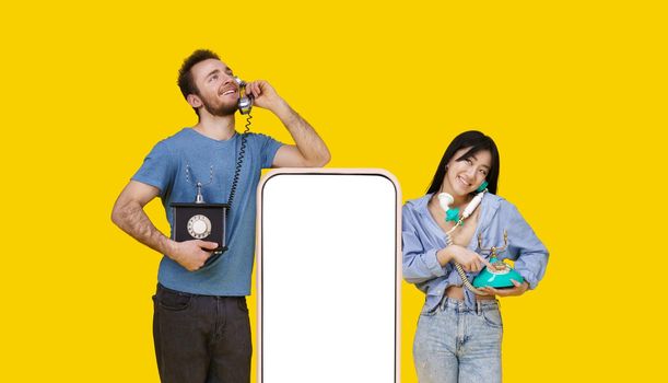 Asian girl and caucasian guy in love talking on vintage phones leaned on huge smartphone with blank screen, looking at each other isolated on yellow background. Product placement.
