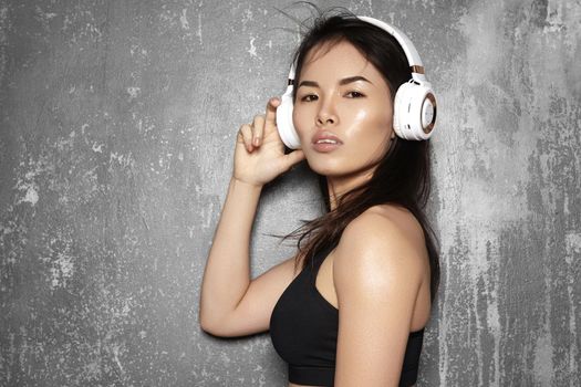 Beautiful sport woman with big white headphones. Model listening the music. Fitness portrait on gray wall. Perfect body shapes