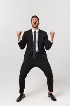 Cheerful young bearded business man show hand up excited with clenched fists. Full length portrait business man isolated over white studio background.