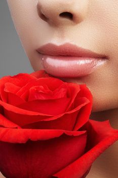 Close-up beautiful female lips with bright lipgloss makeup. Perfect clean skin, light fresh lip make-up. Beautiful spa portrait with red rose flower. Spa and cosmetics