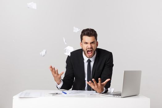 Angry young bearded man work at desk with laptop isolated over white background. Screaming tearing paper documents.