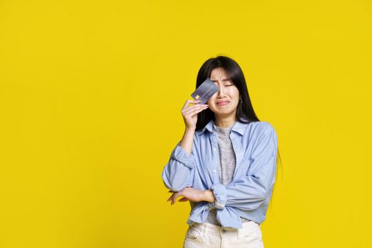 crying unhappy asian girl holding dark credit, debit card touching her eye hiding begging for money. Asian girl in blue shirt with mockup credit card isolated on yellow background. Shopping concept.