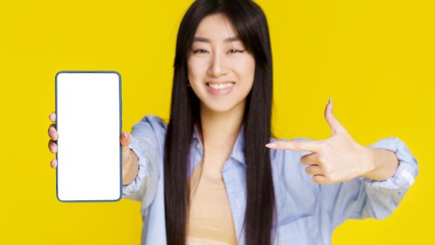 Mobile phone app advertisement. Young asian girl happy holding smartphone pointing on white empty screen and exciting to win isolated on yellow background. Product placement. Selective focus on phone.