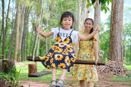 Young mother swings for her daughter in the park. Smiling little girl sitting on swing under tree with mother in summer park. Happy family having fun in the park.