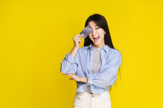 Happy asian girl holding credit, debit card touching her face, eye. Asian girl in blue shirt holding mockup credit card isolated on yellow background. Shopping concept.