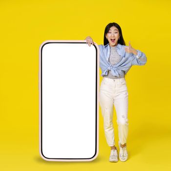 Excited asian girl gesturing thumb up leaned on giant, huge smartphone with white, blank screen standing leaned on wearing casual isolated on yellow background. Free space mock up.