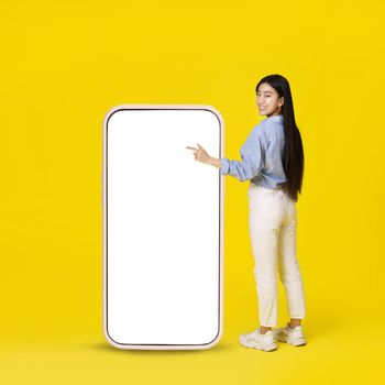 Asian girl pointing on giant, huge smartphone with white blank screen standing back happy pointing finger up dressed in casual isolated on yellow background. Free space mock up.