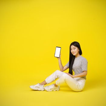 Charming asian young girl holding smartphone sitting on the floor showing a white screen isolated on yellow background. Great offer. Product placement. Mobile app advertisement. Copy space.