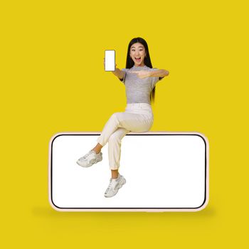 Asian young girl holding smartphone sitting on giant, huge smartphone with black screen isolated on yellow background. Mock up product placement. App Advertisement. Copy space.