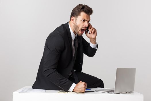 Angry businessman sitting at the table and screaming over gray background. Looking at camera.