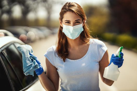 A young woman with a mask on her face and protective gloves on her hands is ready to wipe her car holding a cloth in one and a bottle with disinfectant in the other hand.