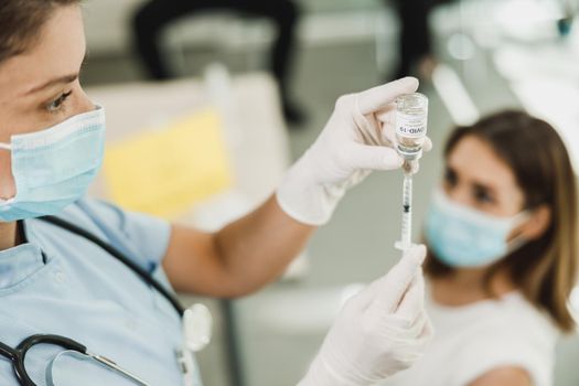 Nurse holding syringe and making Covid-19 vaccination injection dose in shoulder of female patient wearing mask.