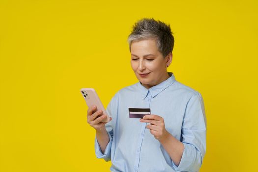 Mature grey haired woman hold smartphone and debit or credit card in hand making online payment or shopping. Pretty woman in blue shirt buying online isolated on yellow background. Focus on the phone.
