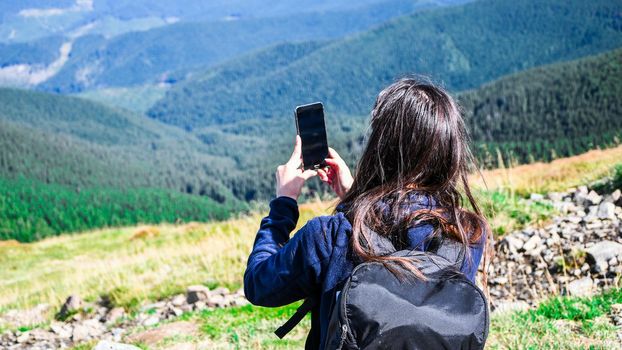 Faceless girl taking photos in mountains. Back view of woman blogger in mountain hiking. Tourist traveler girl in an adventure traveling on the mountains, using phone taking a photograph of the misty mountain view