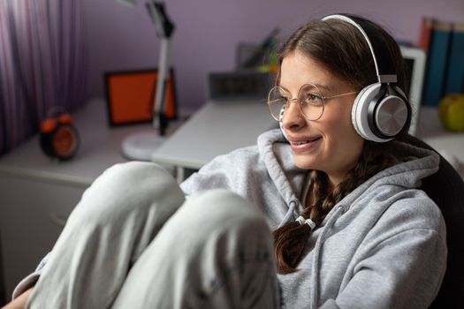 Brunette girl with glasses sits at her room and listens to music. A young girl smiling and full of musical excitement.