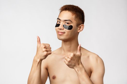 Beauty, people and lifestyle concept. Portrait of sassy good-looking asian man with naked torso, show thumbs-up in approval, use eye-patches for skincare routine, smiling satisfied, white background.