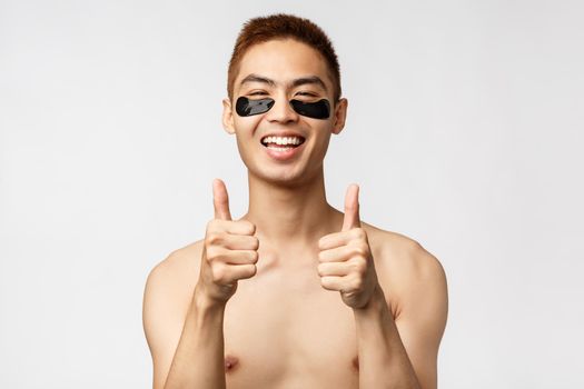 Beauty, people and lifestyle concept. Handsome asian man with naked torso, looking satisfied, self-distancing during codi19 quarantine and take care of skin, apply eye-patches, white background.