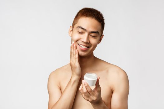 Beauty, people and leisure concept. Happy, silly handsome asian naked man close eyes, apply daily cream, smiling delighted as taking care of acne prone skin, standing white background.