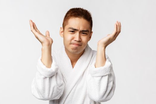 Beauty, spa and leisure concept. Portrait of distressed, pissed-off asian man in bathrobe, pouting troubled and raise hands unaware, feel frustrated and confused, grimacing bothered.