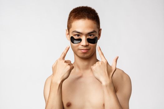 Beauty, people and lifestyle concept. Portrait of young asian man with naked torso, wearing black eye-patched to take care of skin, pointing fingers at face and look camera, white background.