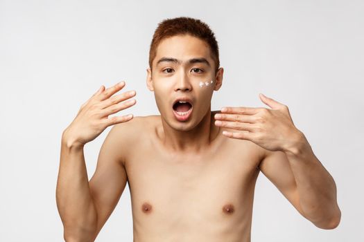 Beauty, people and leisure concept. Portrait of charismatic asian man with naked torso, shaking hands and gasping astonished, react to something overwhelmed, apply skincare cream under eyes.