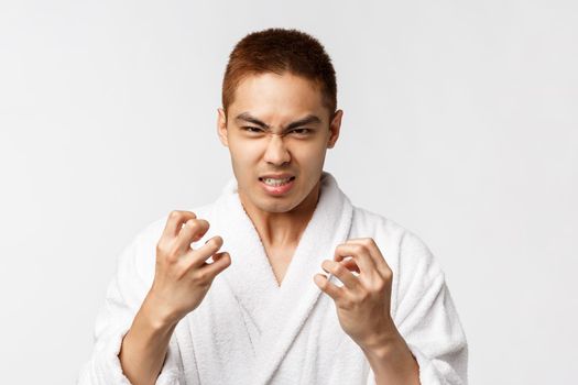 Beauty, spa and leisure concept. Portrait of angry, hateful asian man in bathrobe look with hate and anger camera, clench hands into fists, feel aggression, standing white background annoyed.