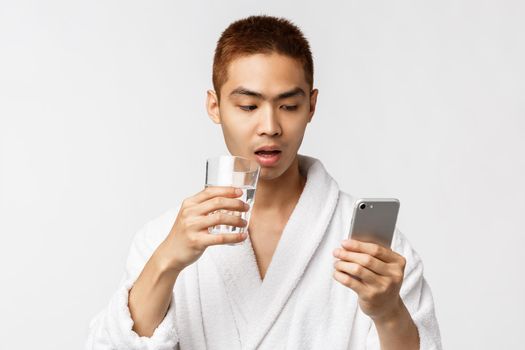 Beauty, spa and technology concept. Portrait of curious and amazed asian man in bathrobe, drinking water from glass and capture attention by interesting news on mobile phone, white background.