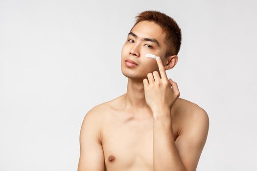 Beauty, people and leisure concept. Sassy good-looking asian man with naked torso taking care of his perfect, no blemishes skin, apply cream on face and look camera pleased, white background.