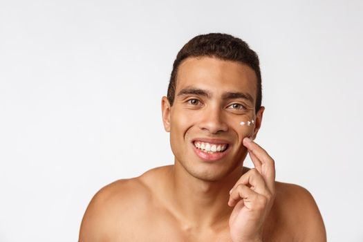 Photo of shirtless african american man smiling and applying face cream isolated over white background.