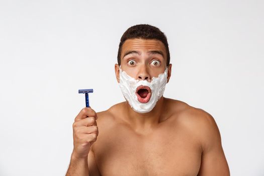 Close up of a happy naked african man with face in shaving foam holding razor isolated over gray background