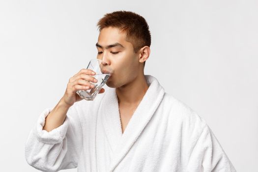 Beauty, spa and technology concept. Portrait of young handsome healthy asian man, drinking water from glass and wear bathrobe, standing white background, start morning with sip of aqua.