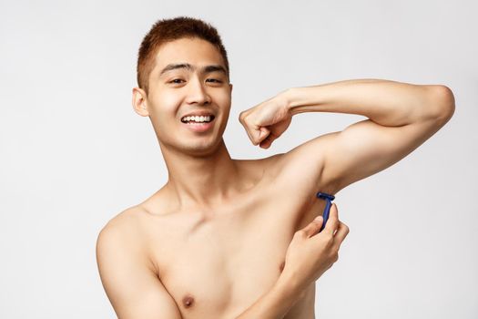 Beauty, people and hygiene concept. Handsome confident and pleased, smiling asian man standing naked showering, washing armpit with sponge, look camera delighted, white background.