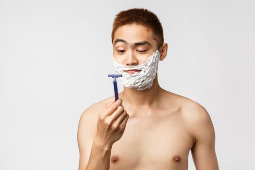 Beauty, people and hygiene concept. Curious handsome asian man looking at new razor as starting shaving beard, standing with cream on face and naked torso, get ready to work, bathroom routine.