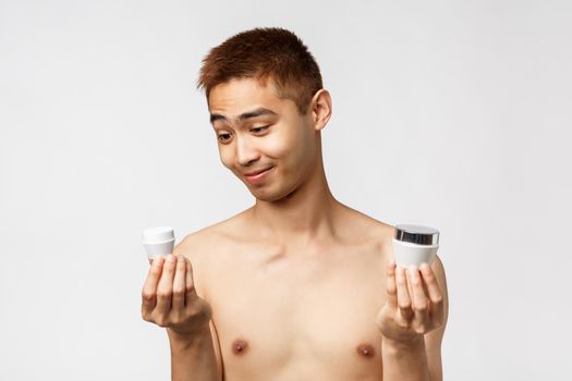 Beauty, people and leisure concept. Pleased smiling asian man with naked torso, smiling as looking at small adorable bottle of cream, apply skincare routing cosmetics product, white background.