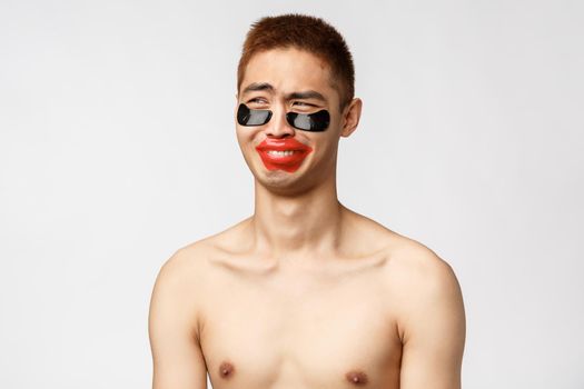 Beauty, people and leisure concept. Funny and silly asian naked boyfriend, laughing and looking away, girlfriend making fun of him applying eye-patches and lip-patch, white background.