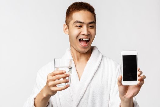 Beauty, spa and leisure concept. Happy young asian man in bathrobe promote smartphone application to check on healthcare, showing mobile phone screen and holding glass with water.