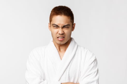 Beauty, spa and leisure concept. Portrait of grumpy, hateful asian man look with disdain and hate, grimacing angry camera, feel offended and annoyed, wear bathrobe, white background.