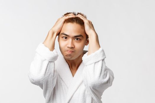 Beauty, spa and leisure concept. Portrait of sick and tired, tensed angry asian man holding hands on head, biting lip and frowning, feel stress, standing frustrated and bothered, white background.