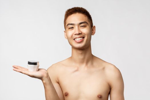 Beauty, people and leisure concept. Pleased good-looking naked asian man with satisfied beaming smile, holding cream jar, recommend skincare routine product, white background.