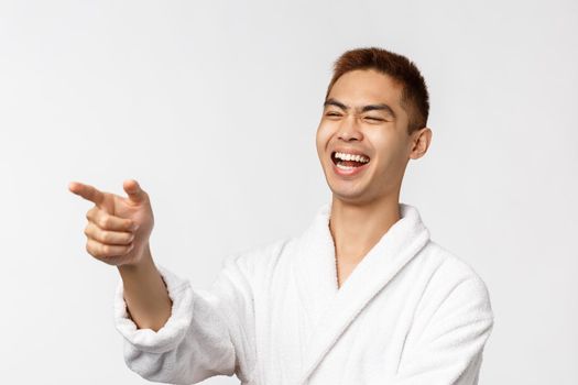 Beauty, spa and leisure concept. Amused, enthusiastic handsome asian man social-distancing, pointing finger left and laughing at something hilarious, watching tv series on quarantine.