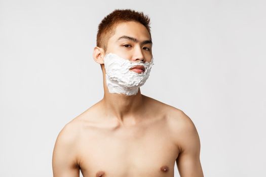 Beauty, people and hygiene concept. Good-looking asian man with naked torso looking at himself in mirror applied shaving cream on face to get rid of beard, standing white background.