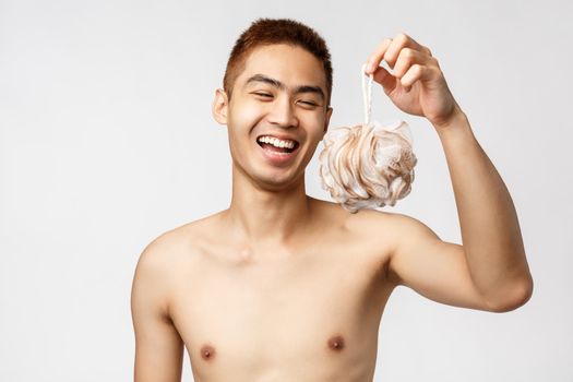 Beauty, people and hygiene concept. Happy young handsome asian man standing naked over white background, holding shower sponge and laughing. showering after sport workout.