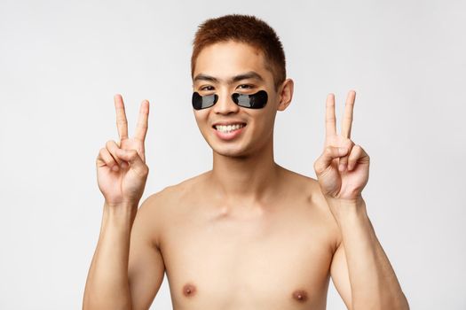 Beauty, people and lifestyle concept. Close-up portrait of happy, relaxed naked asian man wearing eye-patches, show peace signs and smiling, enjoying weekend relax and spa time, skincare routine.